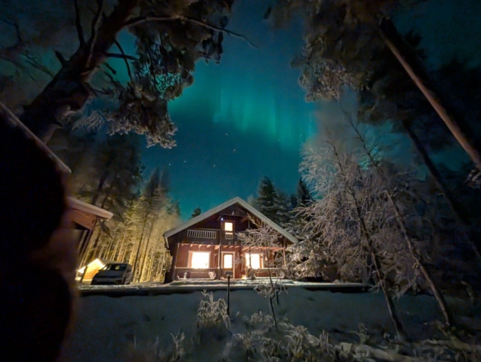 Nordlichter über dem beleuchteten Haupthaus zwischen hohen Bäumen